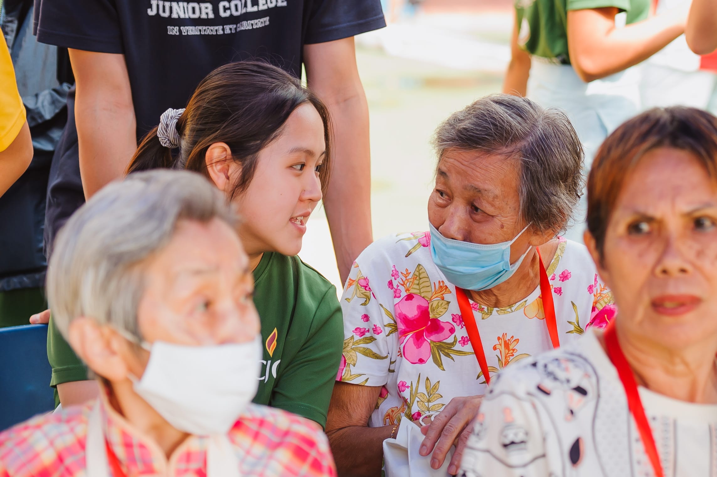 Young woman in green shirt talks with elderly women wearing face masks outdoors during a community gathering.